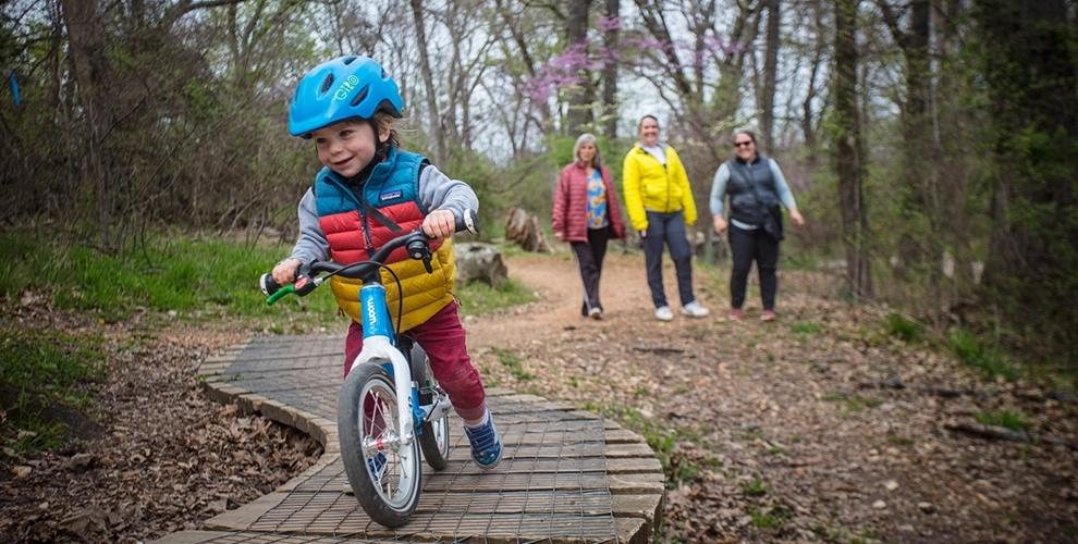 Young kid riding over a wooden feature on a balance bike smiling while family looks forward smiling in the distance.