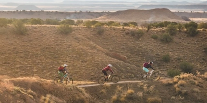Three women riding dirt singletrack from left to right in frame, in desert landscape. 